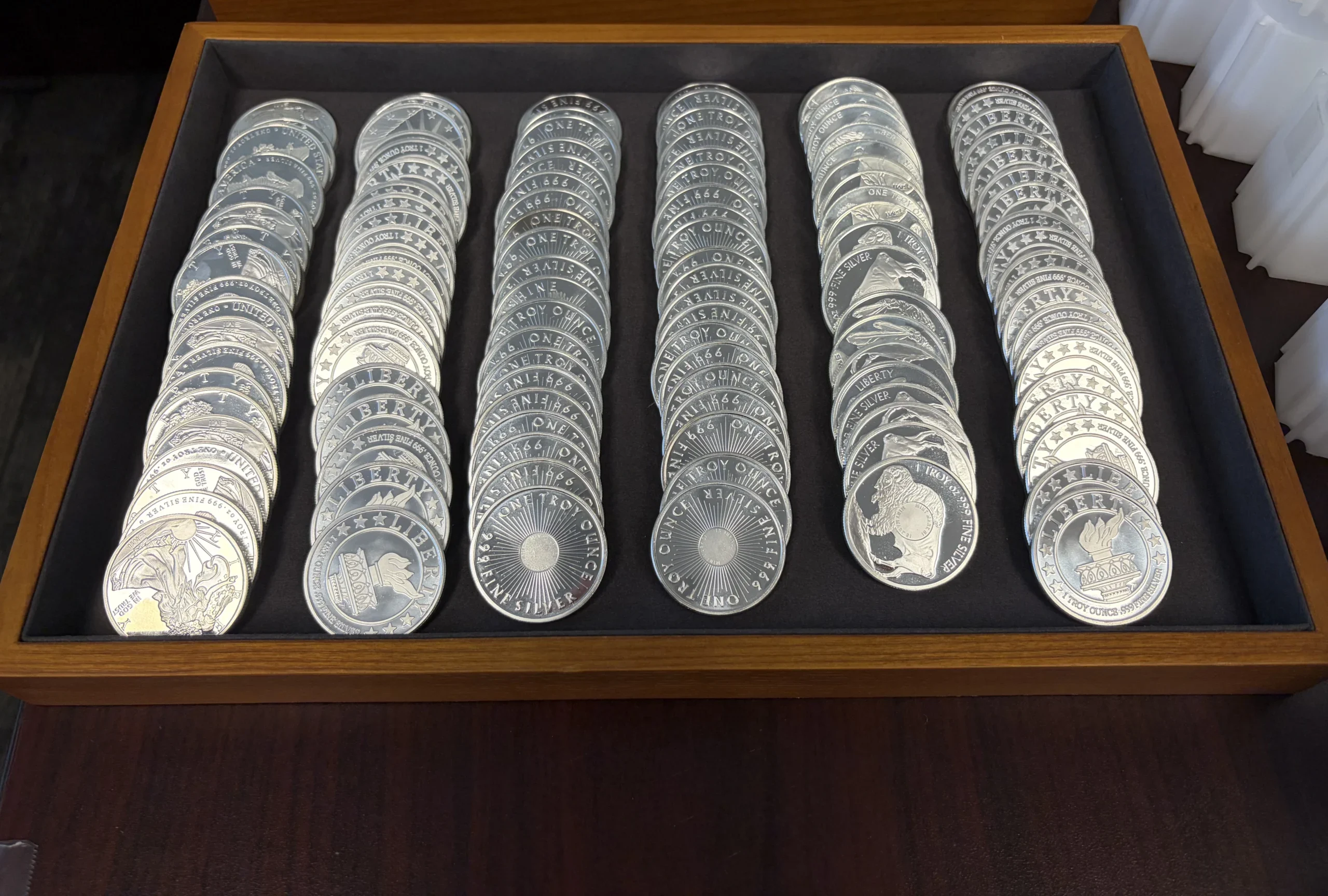 Rows of shiny silver coins arranged in wooden display trays inside a case. | Metals Edge Rows of shiny silver coins arranged monetary in wooden display trays inside a case.