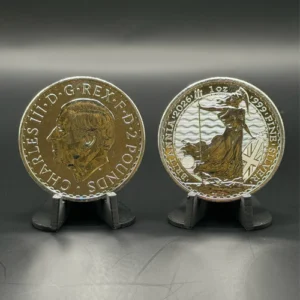 1 oz British Silver Britannia | Metals Edge Two commemorative coins on small display stands, silver with gold-toned centers, shown side by side on a dark surface; left coin features a profiles portrait with surrounding inscriptions, right coin shows a seated figure with a flag and a Union Jack motif.